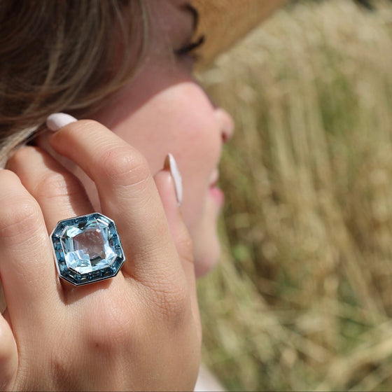 Bague pour femme en or blanc pavée de diamants blancs avec un solitaire d'aigue-marine entouré de topazes bleues. Bague Michaud 143790